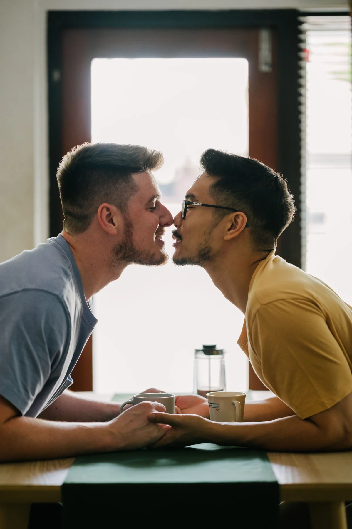 Two men sharing a kiss and holding hands across a cafe table
